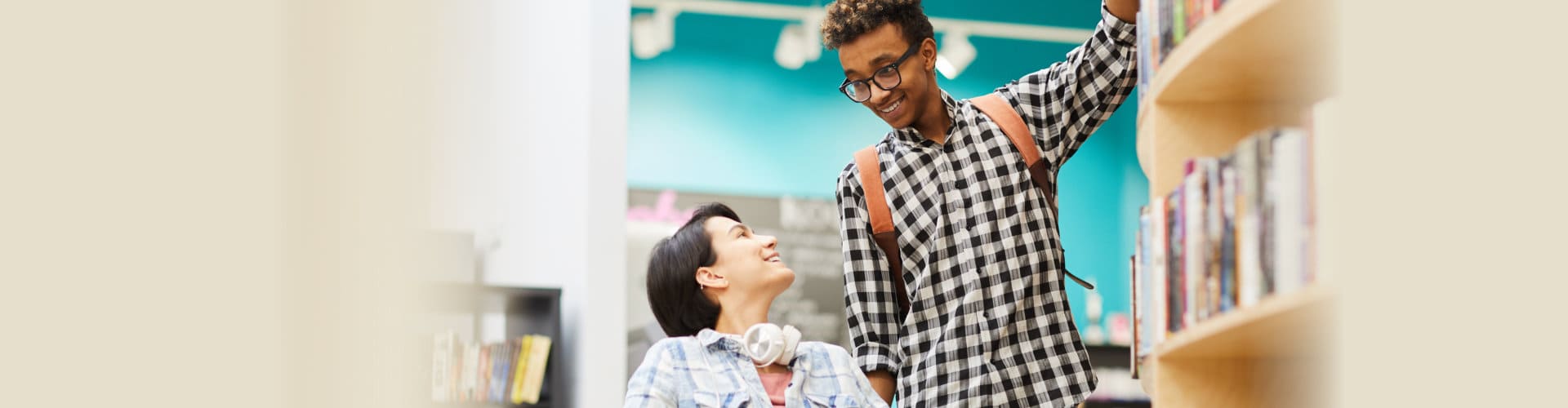 african guy with Afro hairstyle helping disabled student girl to take book from shelf in library